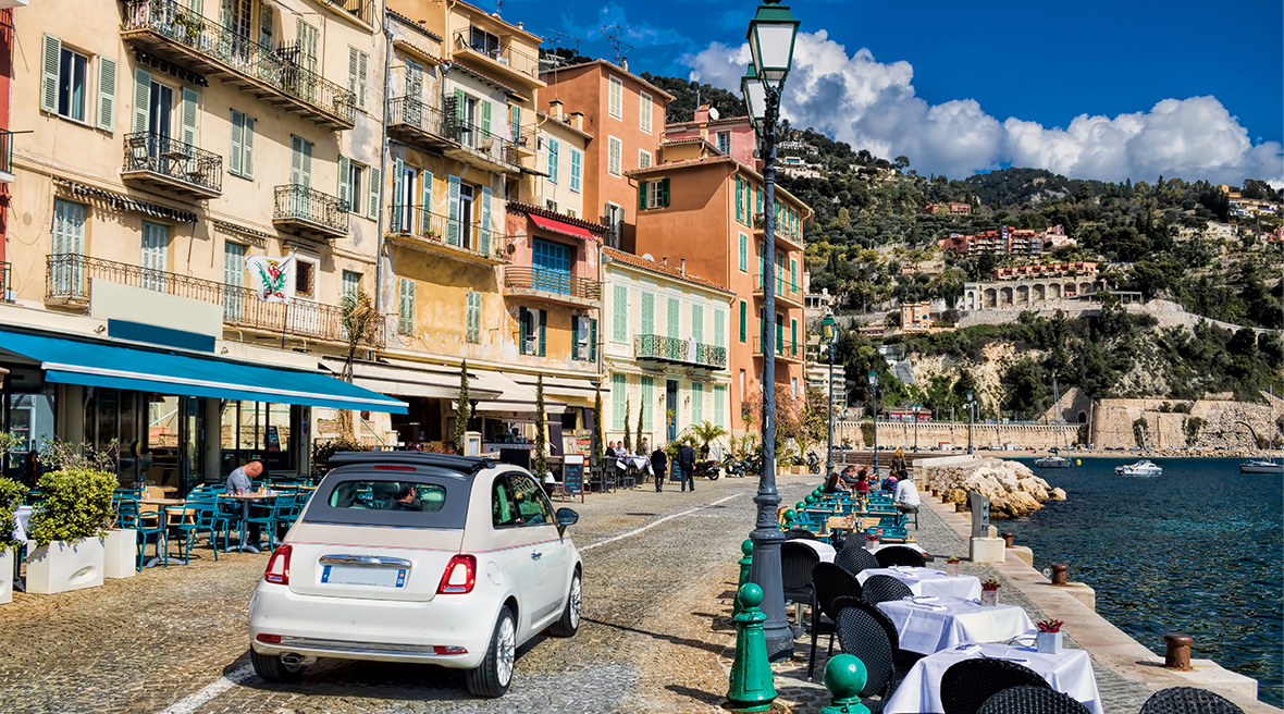 A white Fiat500 driving along the waterfront of a picturesque French town in the sun